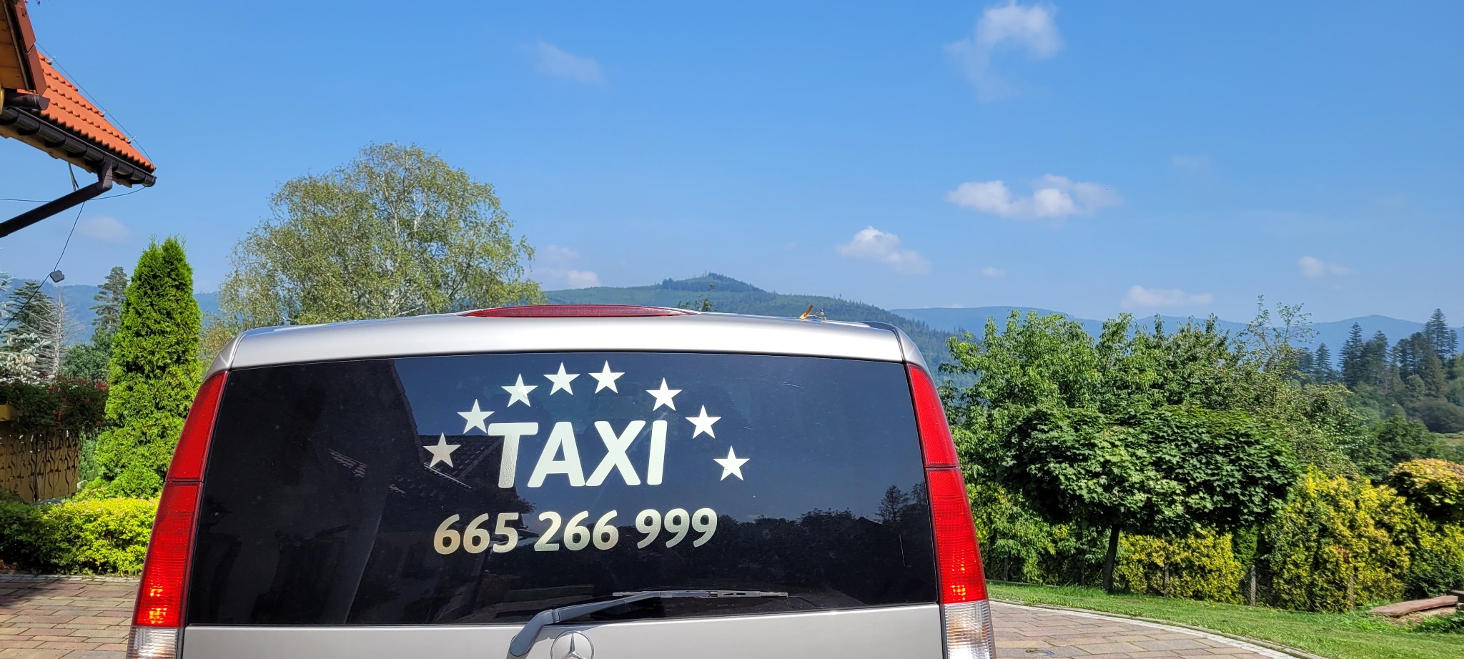 Srebrny samochód typu van z naklejką 'TAXI' otoczoną gwiazdami i numerem telefonu na tylnej szybie, na tle górskiego krajobrazu w słoneczny dzień.