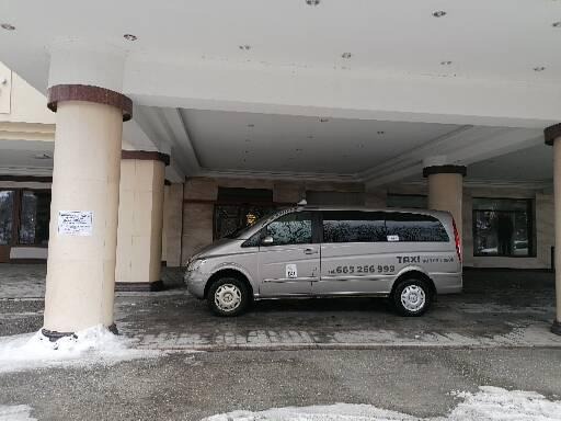 Srebrny minivan z napisem TAXI zaparkowany pod zadaszeniem budynku z kolumnami, widoczny numer telefonu 665 266 999.