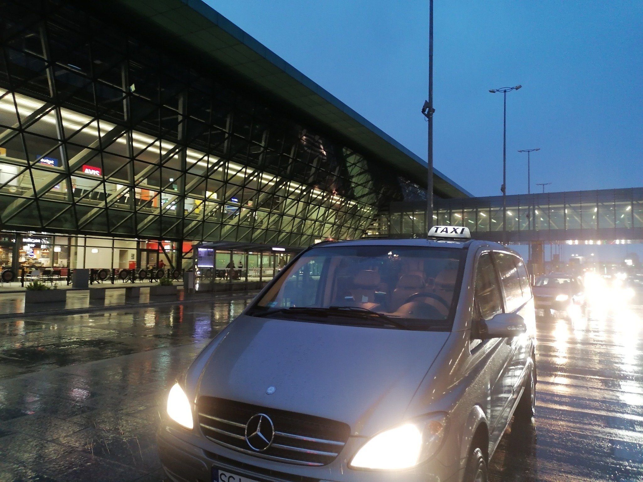Srebrny van Mercedes z napisem TAXI na dachu, stojący na mokrym asfalcie przed nowoczesnym budynkiem lotniska o zmierzchu.