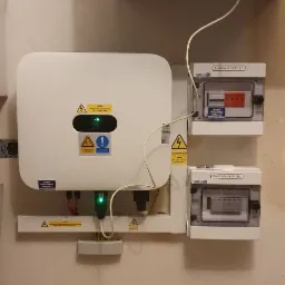 Biały falownik z zieloną diodą i tabliczkami ostrzegawczymi, zamontowany na ścianie obok rozdzielnic PV-AC i PV-DC, widoczne okablowanie i logo firmy Smart PV Group w górnym rogu.