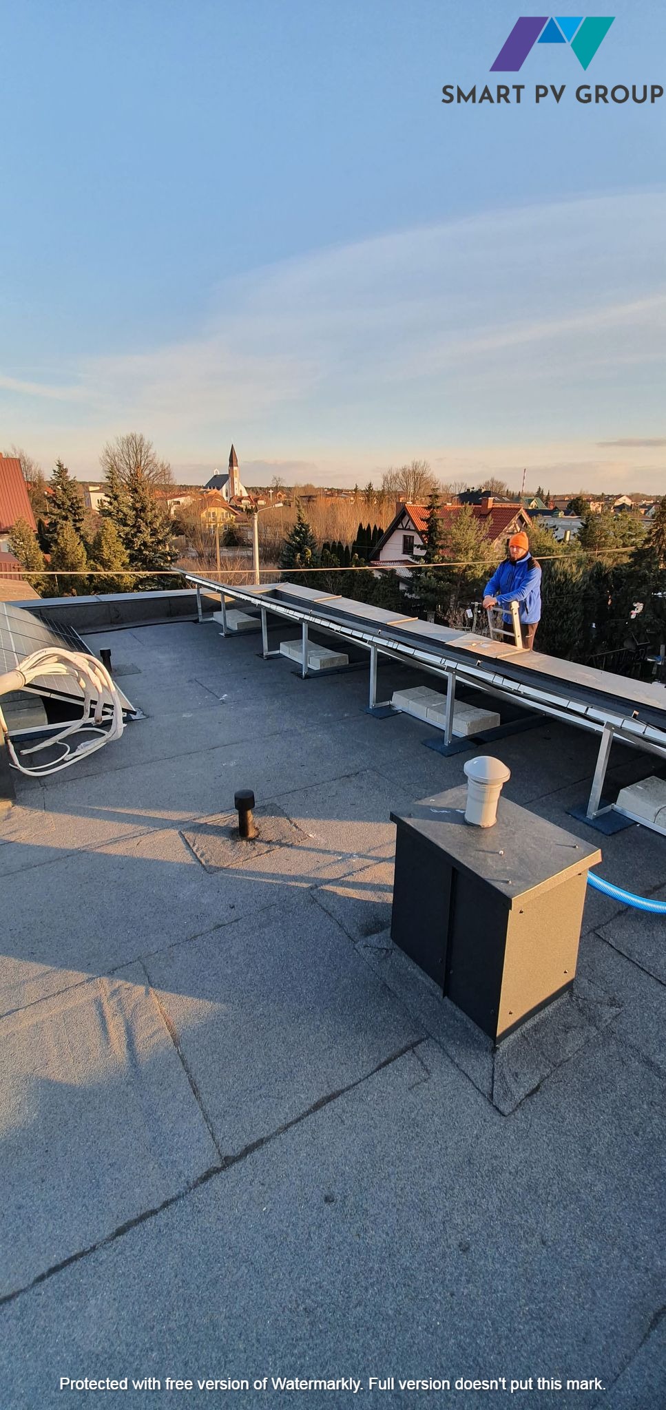 Instalator w pomarańczowym kasku montuje konstrukcję wsporczą na dachu pokrytym papą, w tle zabudowa miejska z kościołem.