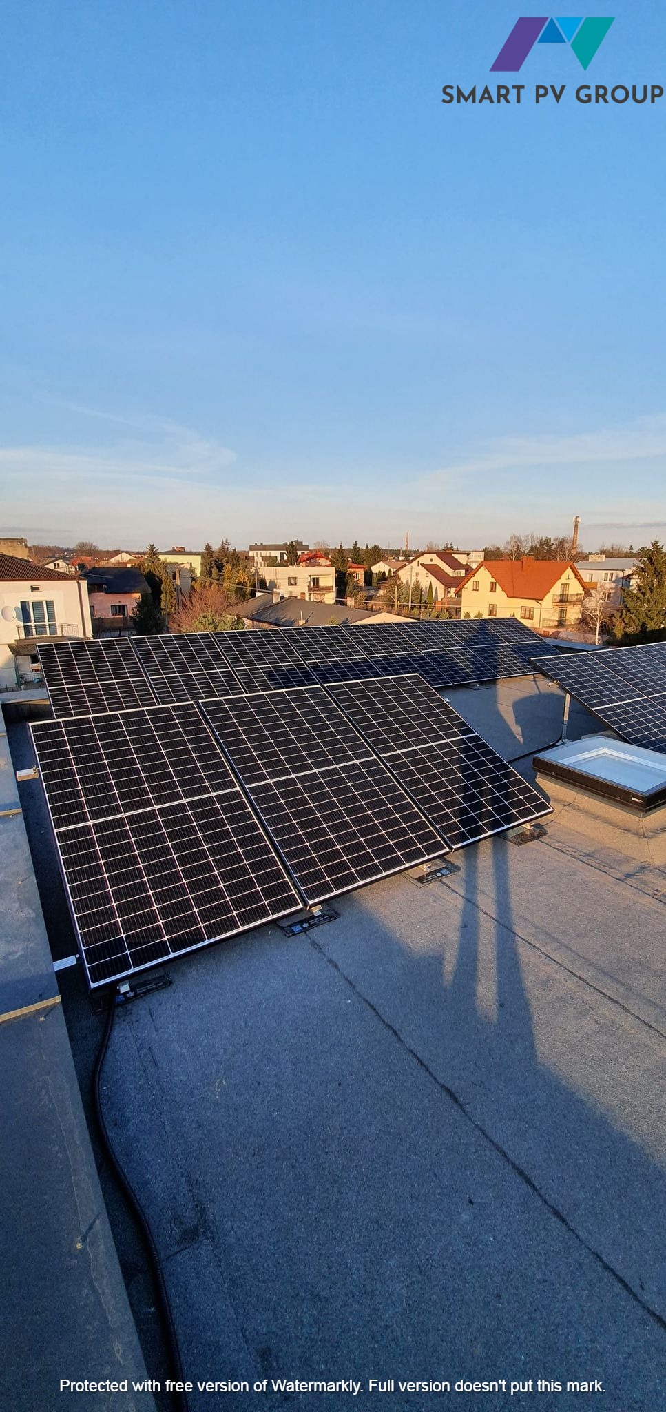 Instalacja paneli fotowoltaicznych na płaskim dachu budynku mieszkalnego, widok z góry na panele w regularnym układzie, w tle zabudowa jednorodzinna i błękitne niebo, logo Smart PV Group w górnej...