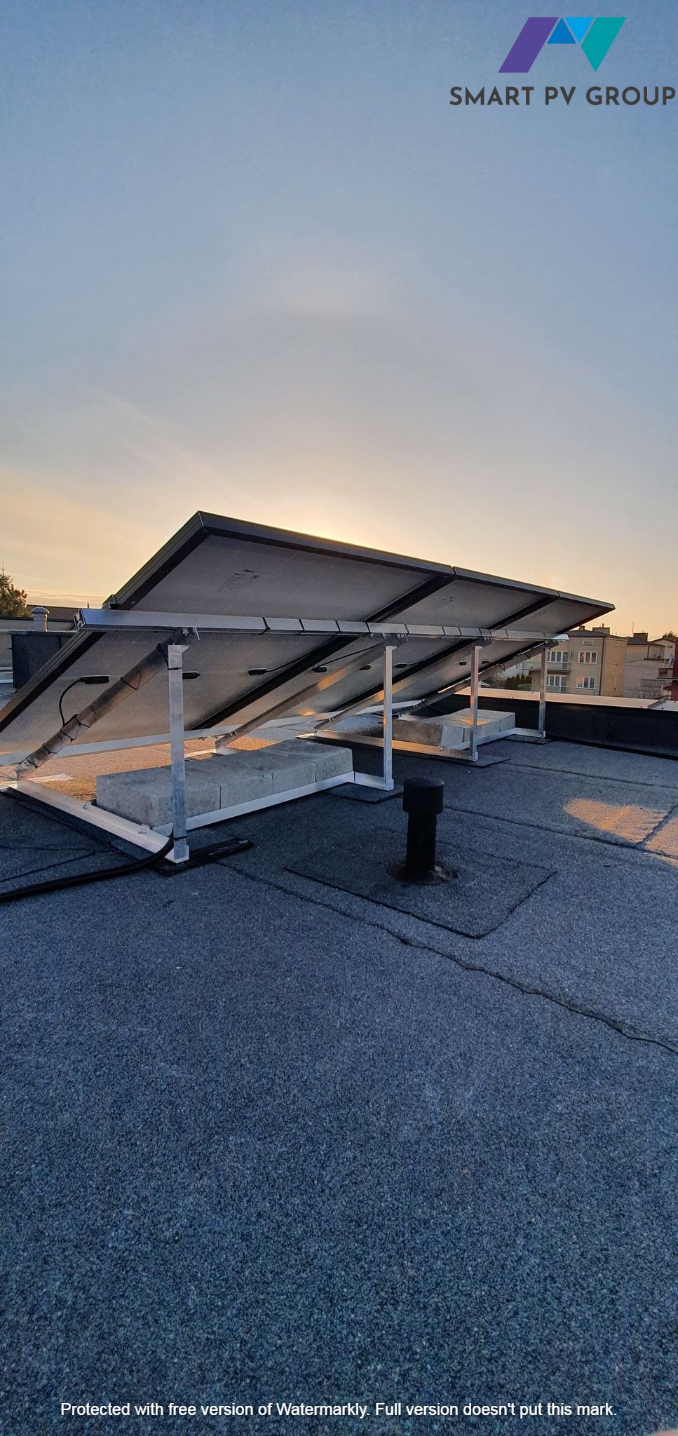 Panele słoneczne zamontowane na płaskim dachu budynku mieszkalnego, widok z dołu na konstrukcję wsporczą i betonowe bloki obciążające, zachodzące słońce w tle.