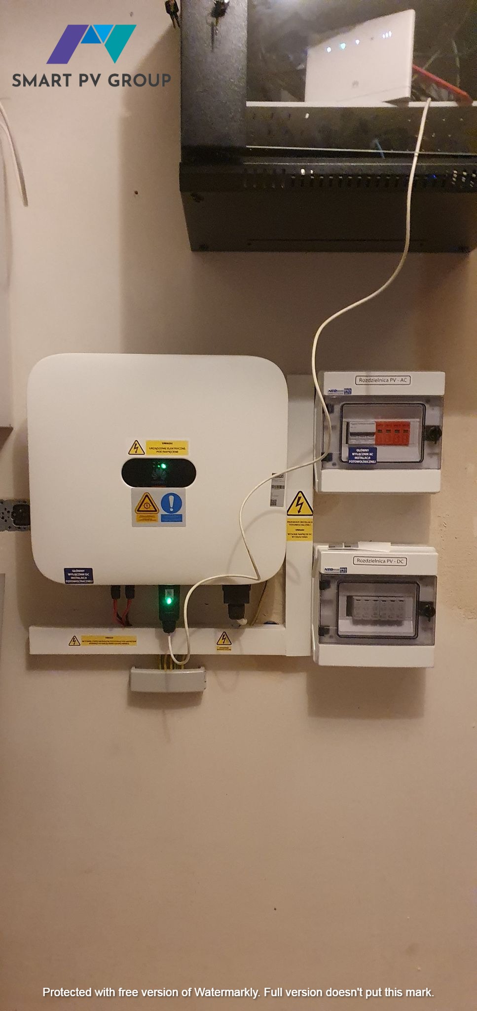Biały falownik z zieloną diodą i tabliczkami ostrzegawczymi, zamontowany na ścianie obok rozdzielnic PV-AC i PV-DC, widoczne okablowanie i logo firmy Smart PV Group w górnym rogu.