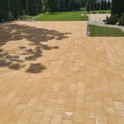 Ułożona, beżowa kostka brukowa na podjeździe przed domem, z widocznym cieniem drzewa i zielonym trawnikiem w tle.