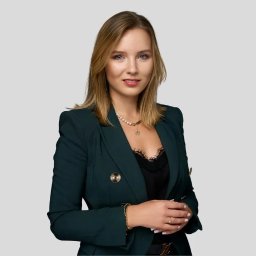 Westate Śródmieście - Portret uśmiechniętej blondynki w ciemnozielonym żakiecie i biżuterii, na jednolitym szarym tle.