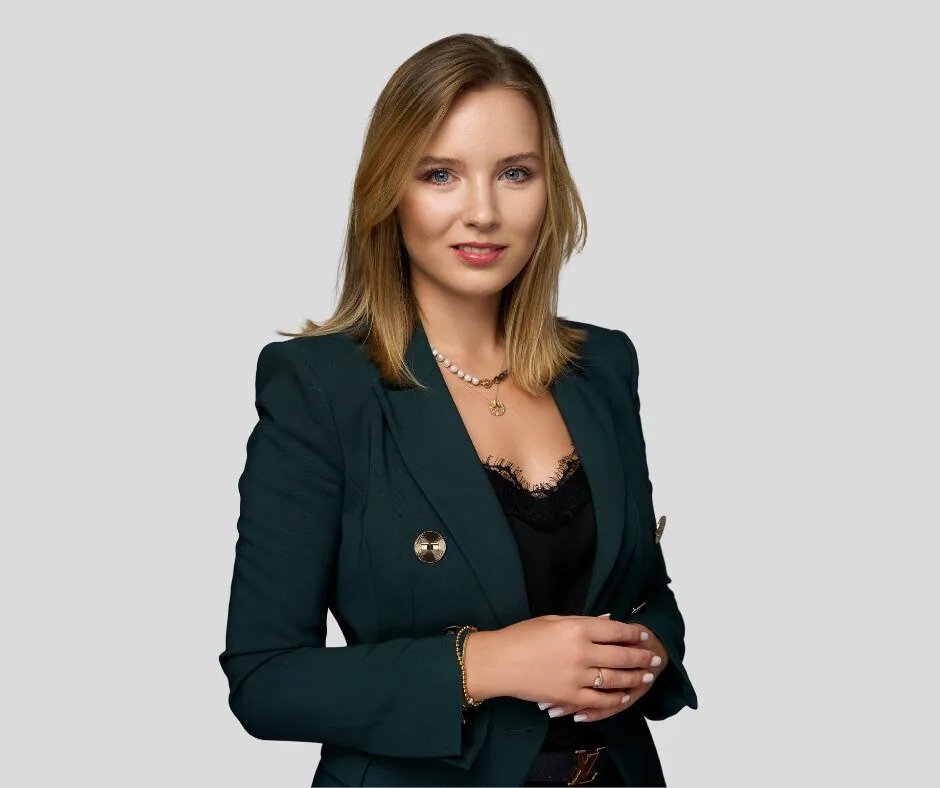 Portret uśmiechniętej blondynki w ciemnozielonym żakiecie i biżuterii, na jednolitym szarym tle.