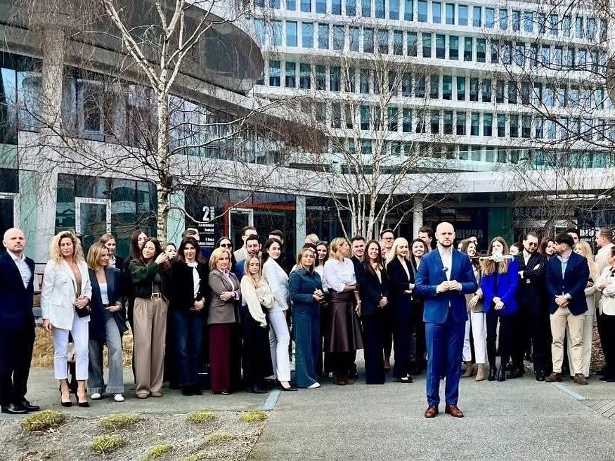 Grupa agentów nieruchomości pozuje na tle nowoczesnego biurowca z logo firmy na elewacji.