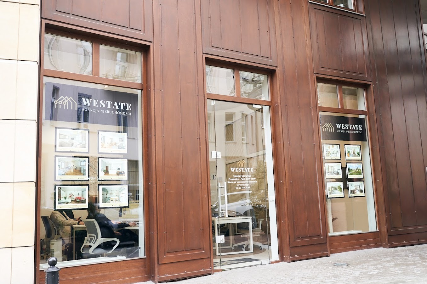 Fasada agencji nieruchomości Westate z brązowymi panelami, widoczne okna z ofertami mieszkań i biuro z pracownikami w środku.