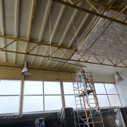 RECOLTE SPÓŁKA Z OGRANICZONĄ ODPOWIEDZIALNOŚCIĄ - Pracownik w kombinezonie ochronnym na rusztowaniu natryskuje pianę izolacyjną na sufit hali z widoczną konstrukcją stalową i lampami przemysłowymi.