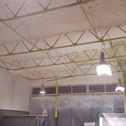 RECOLTE SPÓŁKA Z OGRANICZONĄ ODPOWIEDZIALNOŚCIĄ - Aplikacja izolacji pianką na suficie hali z widoczną konstrukcją stalową, lampami i pracownikiem w kombinezonie ochronnym na rusztowaniu.