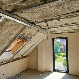 RECOLTE SPÓŁKA Z OGRANICZONĄ ODPOWIEDZIALNOŚCIĄ - Poddasze po ociepleniu pianką PUR, widoczne metalowe profile, okno dachowe i okno pionowe z widokiem na ogród, ściany w trakcie wykańczania.