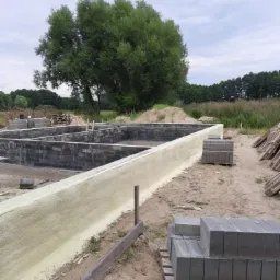 Świeżo zaizolowane fundamenty budynku z widoczną warstwą izolacyjną i paletą z bloczkami fundamentowymi na piaszczystym podłożu, w tle drzewa i pochmurne niebo.