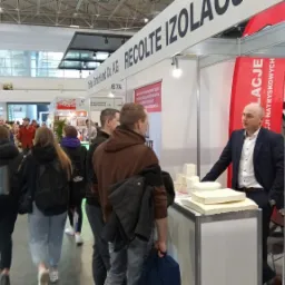 Stoisko targowe firmy izolacyjnej Recoltte Izola z próbkami materiałów izolacyjnych, widoczny mężczyzna w garniturze oraz zwiedzający targi.