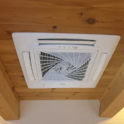 Kasetonowa jednostka klimatyzacyjna Haier zamontowana na drewnianym suficie podwieszanym z widocznymi belkami, widok od dołu.