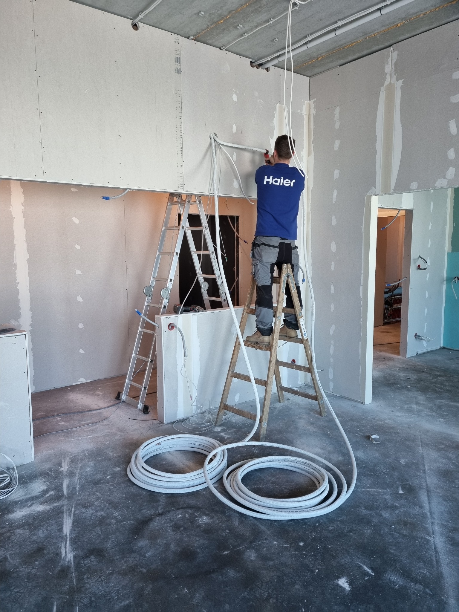 Instalator w niebieskiej koszulce z logo 'Haier' na drabinie, montuje rury w surowym wnętrzu z płyt kartonowo-gipsowych, widoczne zwinięte rury na podłodze.