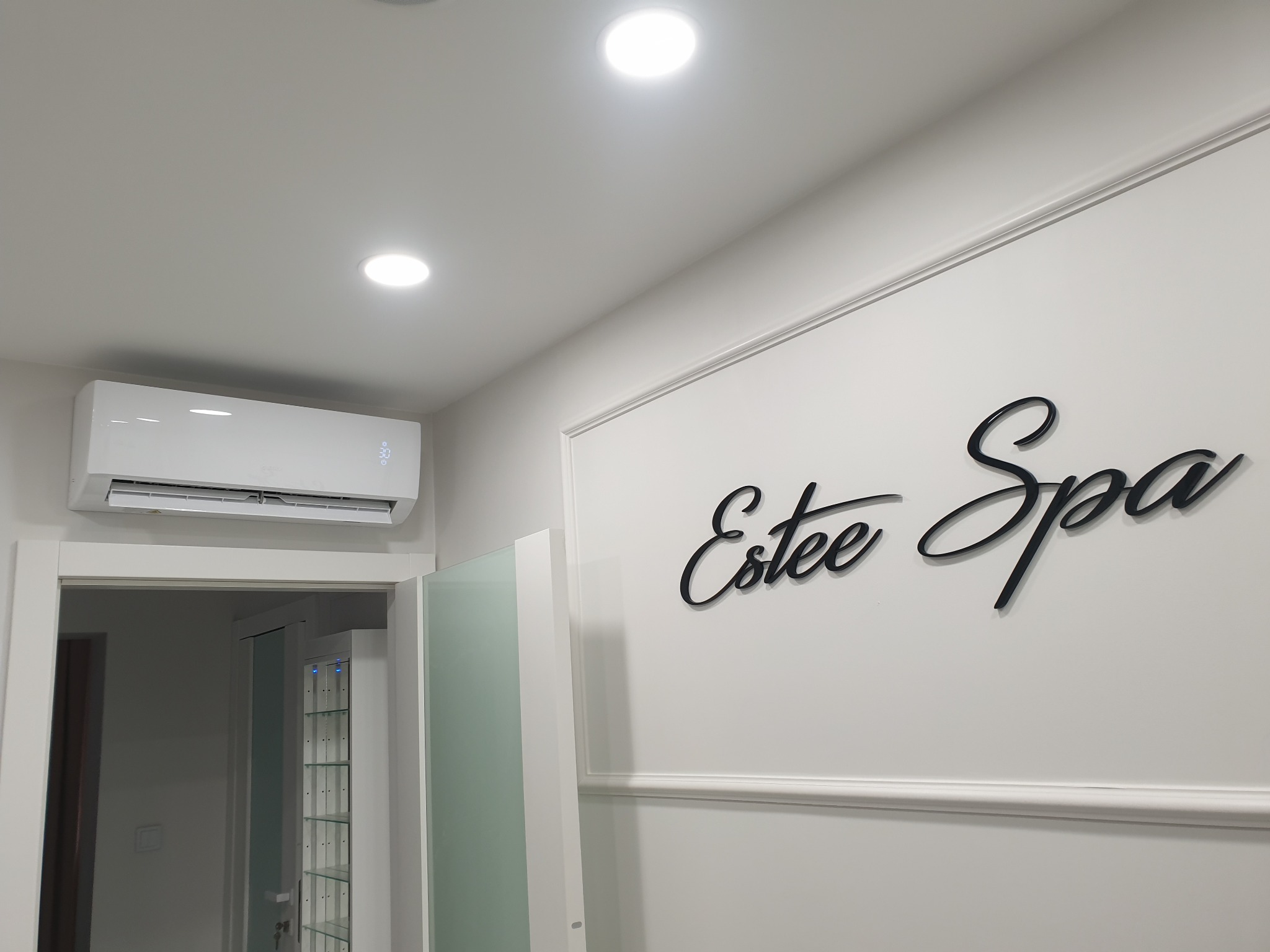 Biała klimatyzacja zamontowana nad drzwiami w minimalistycznym wnętrzu spa z napisem 'Estee Spa' na ścianie.