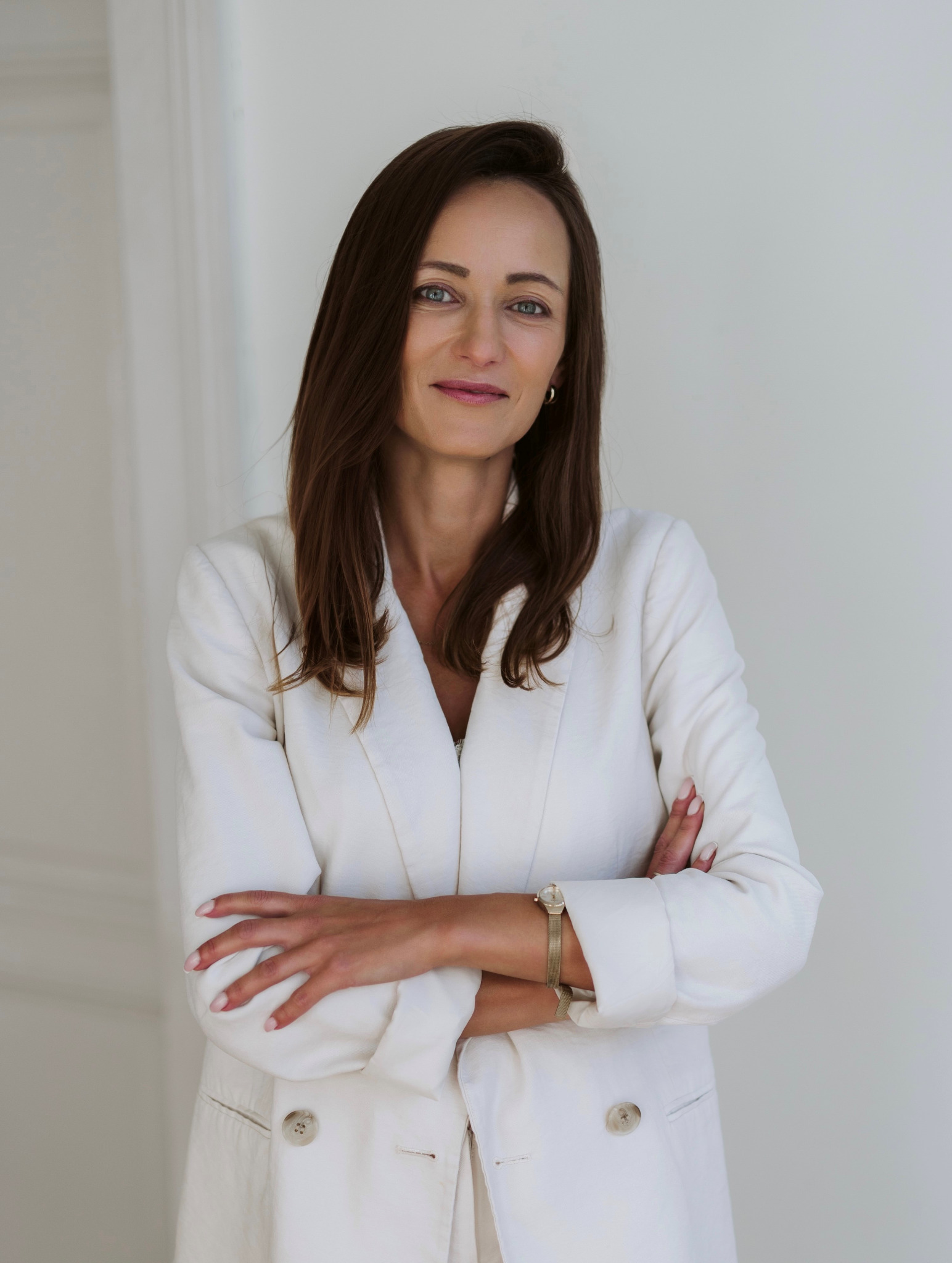 Profesjonalny portret kobiety w białym garniturze z założonymi rękami, zegarek na nadgarstku, na jasnym tle.