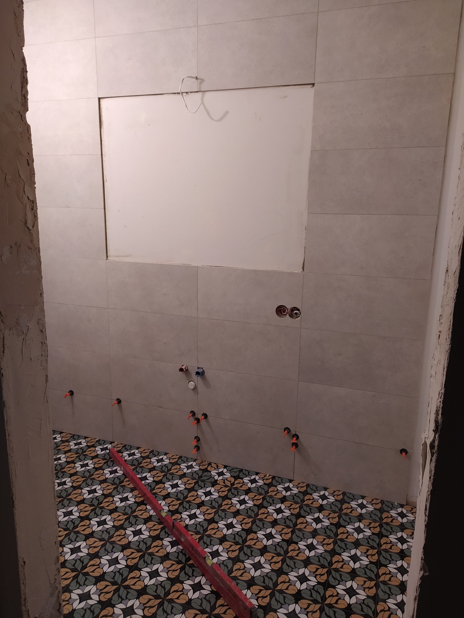 Ściana wyłożona jasnymi płytkami ceramicznymi, z widocznym otworem na wnękę. Na podłodze ułożone płytki z dekoracyjnym wzorem, poziomica leży na podłodze. Widoczne krzyżyki dystansowe na ścianie...