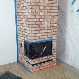 Piotr Waszkiewicz - Wykończony kominek z cegły klinkierowej, zabezpieczony folią i taśmą malarską podczas prac wykończeniowych. Widoczne elementy drewniane i podłoga imitująca drewno.
