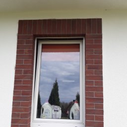 Piotr Waszkiewicz - Otwierane okno z białą ramą, obramowane czerwoną cegłą na białej elewacji. Na parapecie okiennym stoją środki czystości.