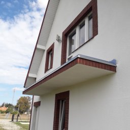 Piotr Waszkiewicz - Elewacja domu po ociepleniu z białym tynkiem i ceglaną obudową okien. Widoczny fragment balkonu. Błękitne niebo w tle.