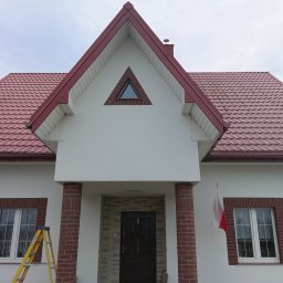 Piotr Waszkiewicz - Biały dom z czerwoną dachówką i ceglanymi detalami, flaga Polski, drabina oparta o ścianę, kot siedzący przed wejściem. Widok z dołu, pochmurne niebo.