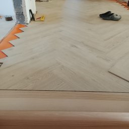 Piotr Waszkiewicz - Układanie jasnych paneli podłogowych w jodełkę, widoczne narzędzia i fragment podkładu. Etap prac wykończeniowych w jasnym wnętrzu.