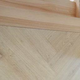 Piotr Waszkiewicz - Jasna podłoga ułożona w jodełkę, z widocznymi drewnianymi listwami przypodłogowymi i fragmentem schodów. Ujęcie z bliska, skupione na detalu wykończenia.