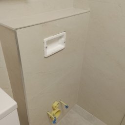 Piotr Waszkiewicz - Nowocześnie wykończona ściana łazienki z zabudową podtynkową WC, jasnymi płytkami, otworem na przycisk spłukujący i wystającymi rurami instalacyjnymi.