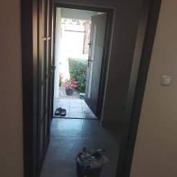 Piotr Waszkiewicz - Ciemna ościeżnica i drzwi wewnętrzne prowadzące na jasne patio z zielonym krzewem i różowymi kwiatami w donicy. Na zewnątrz widoczne klapki, a w przejściu kosz na śmieci.