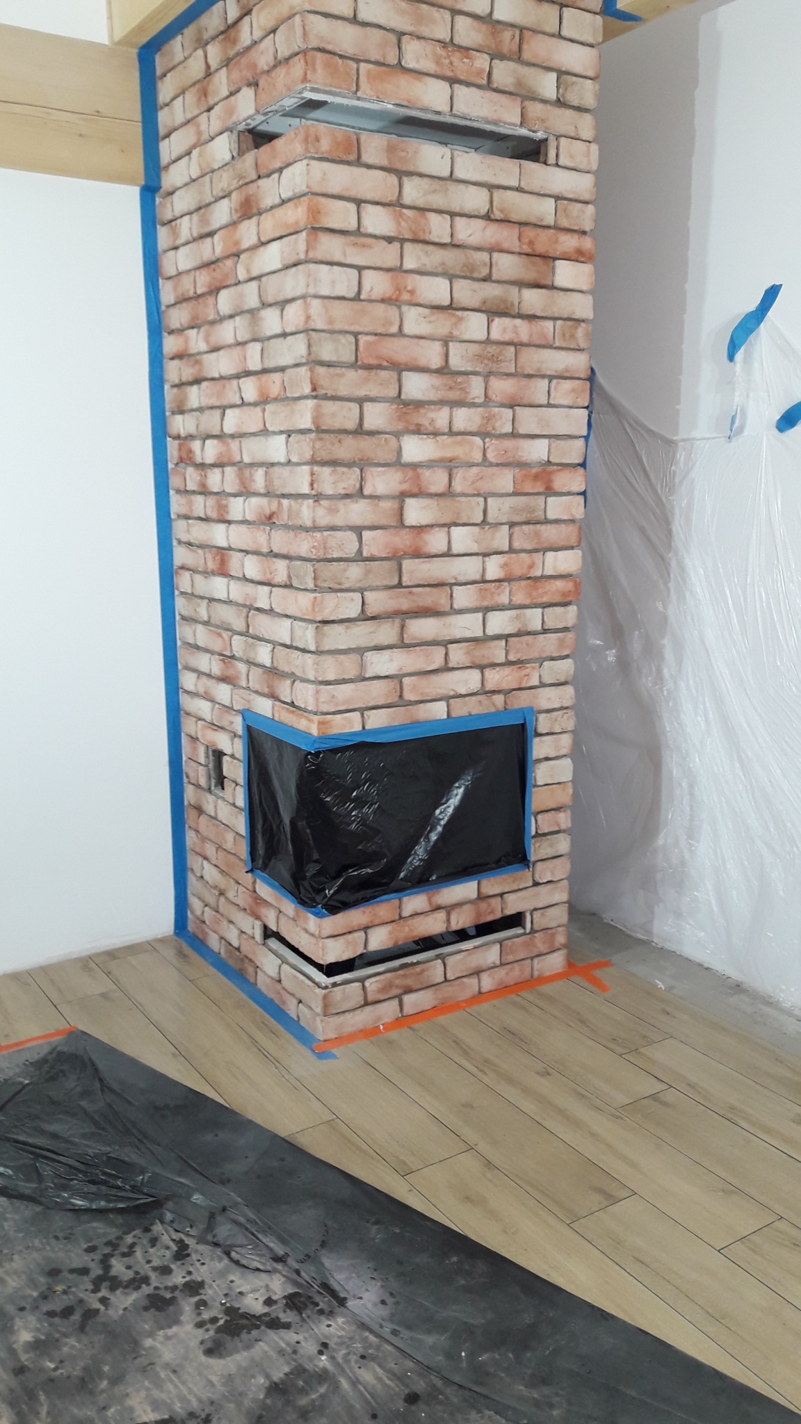 Wykończony kominek z cegły klinkierowej, zabezpieczony folią i taśmą malarską podczas prac wykończeniowych. Widoczne elementy drewniane i podłoga imitująca drewno.