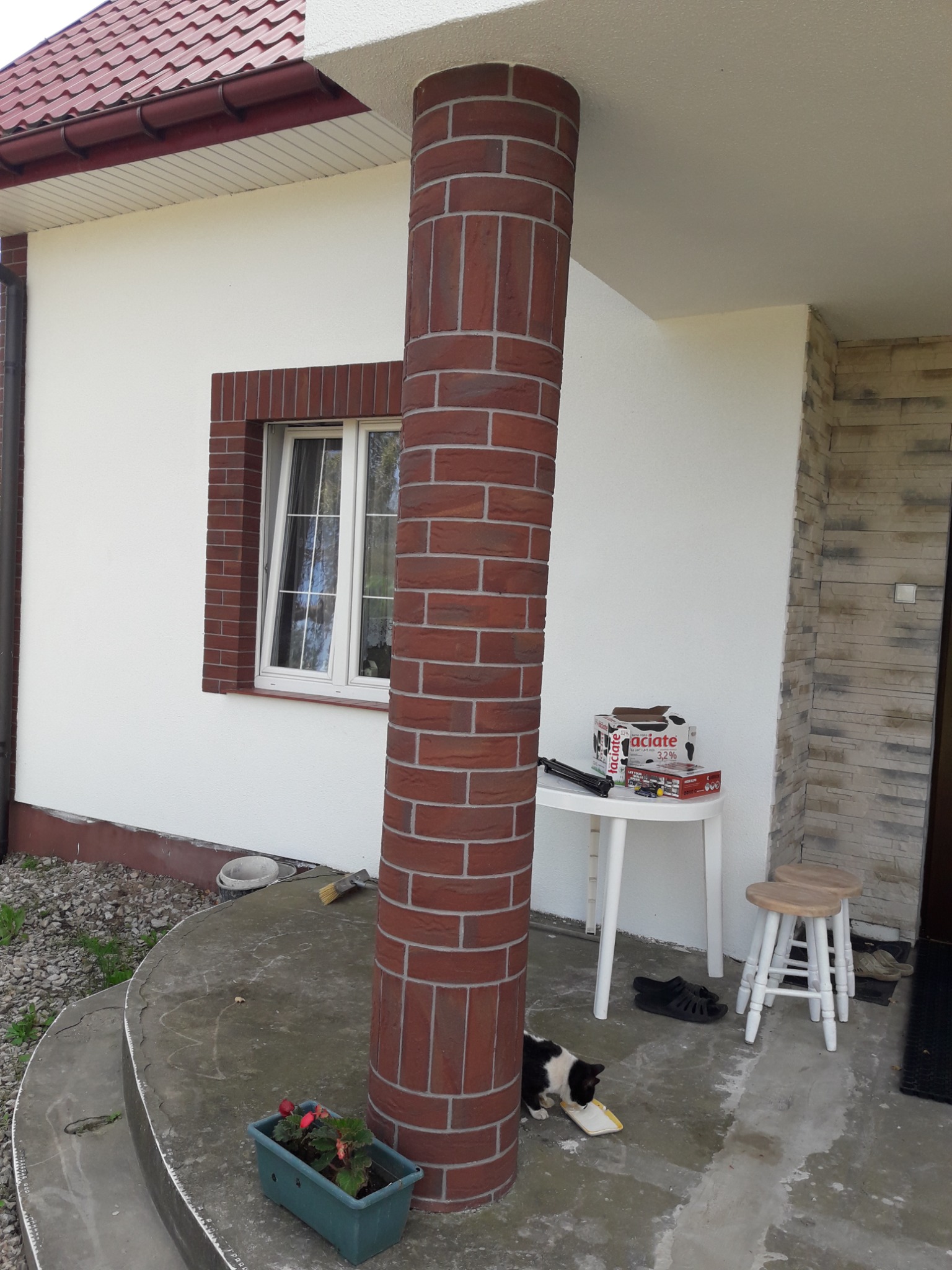 Wykończenie elewacji domu z kolumną imitującą cegłę, okno z ceglaną ramą. Na tarasie kwiaty w doniczce, stolik i stołek. Kot pije mleko z talerzyka.