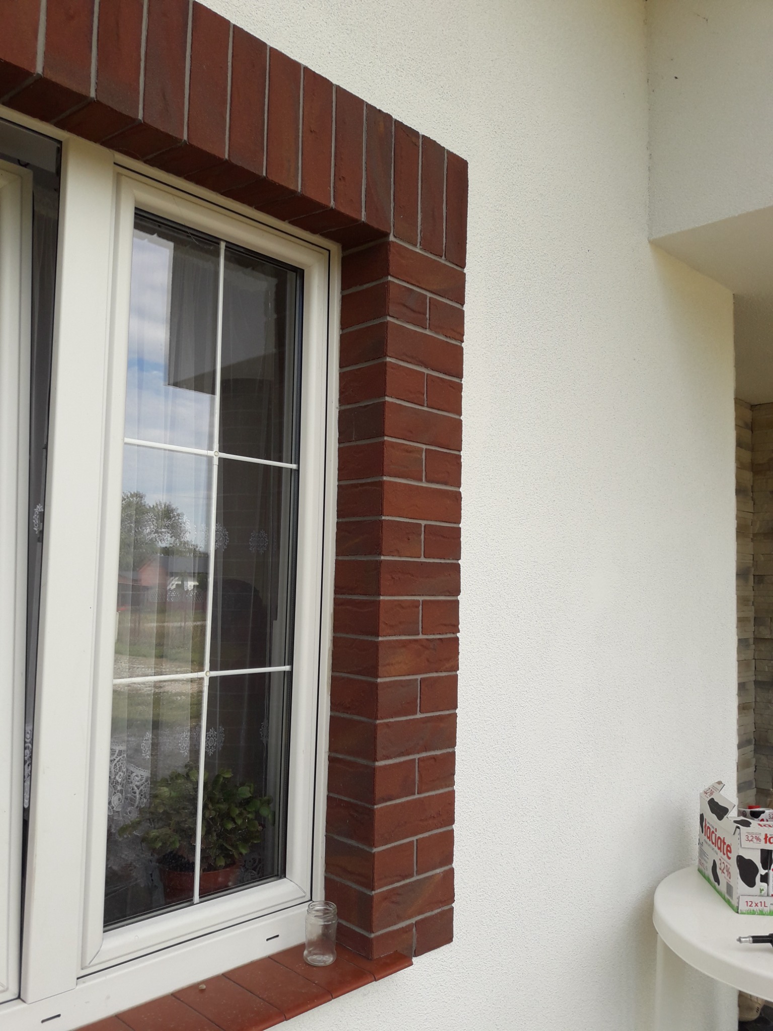 Białe okno PCV z ceglaną obudową, widok z zewnątrz. Dekoracyjna elewacja z białym tynkiem strukturalnym. Na parapecie słoik, w oknie roślina doniczkowa.