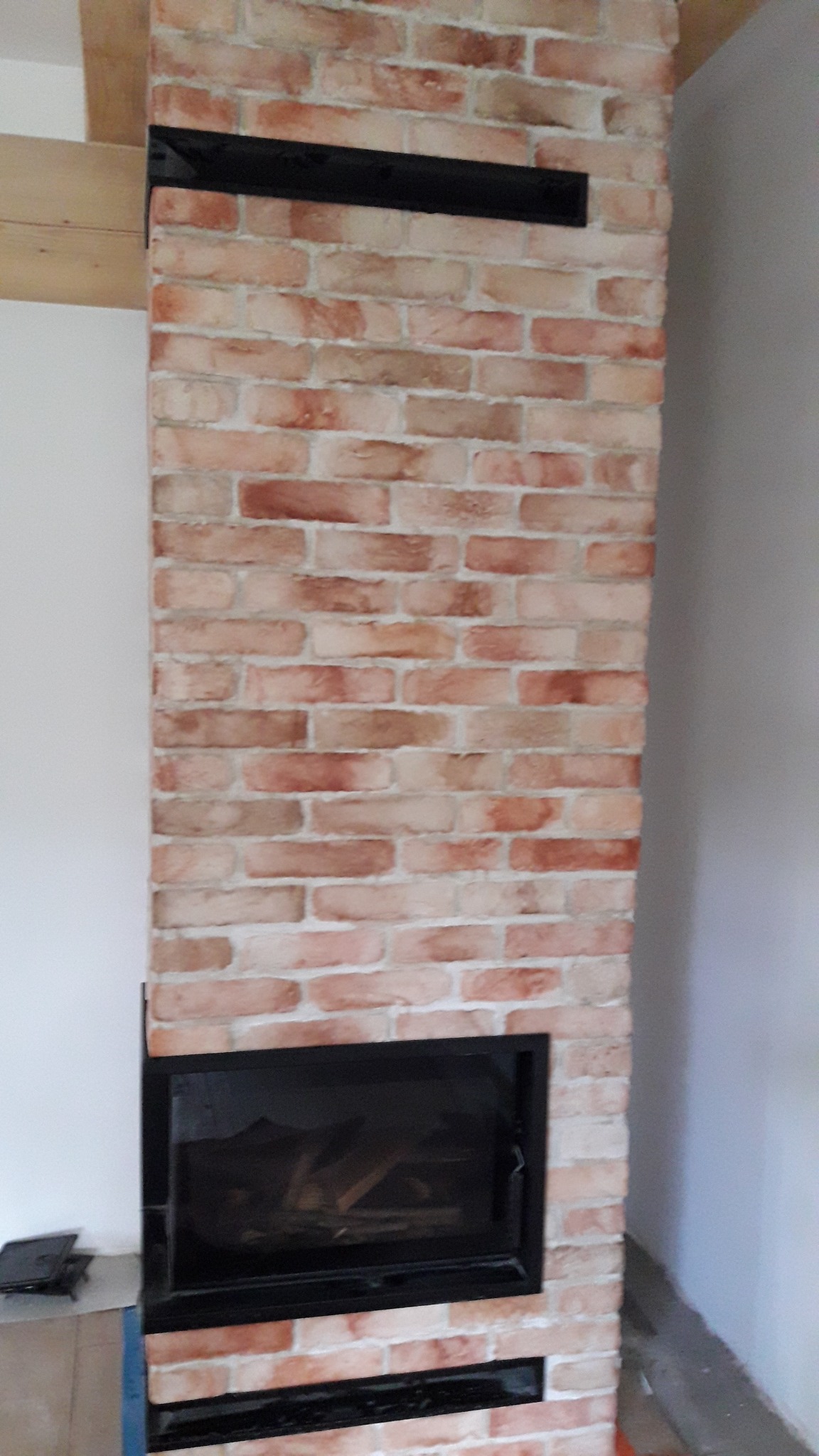 Kominek obudowany cegłą klinkierową z czarnymi wstawkami wentylacyjnymi, widok z dołu. Palenisko zamknięte, z widocznym drewnem w środku. Ściany w odcieniach bieli i szarości.