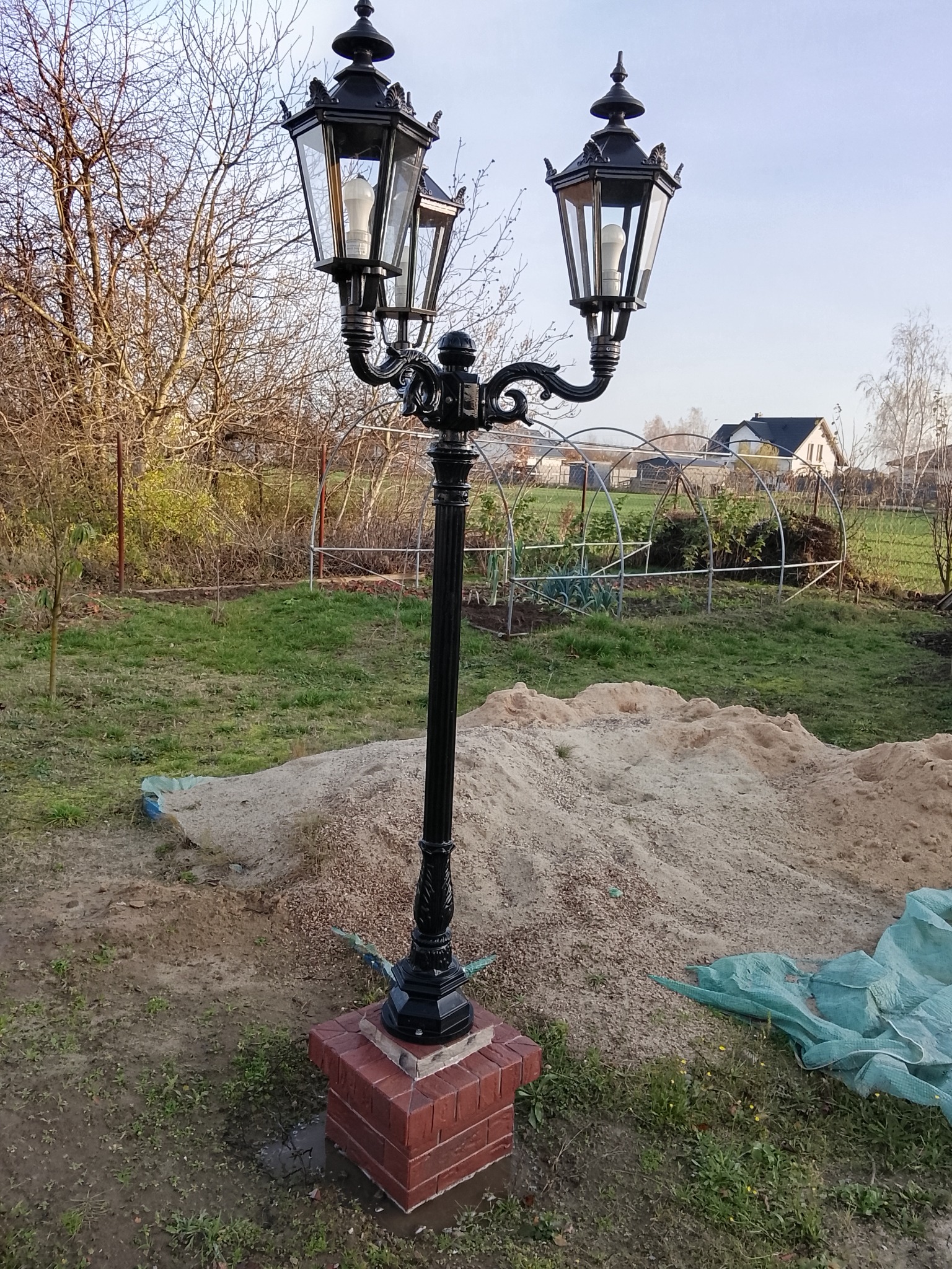 Czarna, ozdobna lampa ogrodowa z trzema kloszami, osadzona na ceglanym cokole. W tle ogród z tunelem foliowym i dom.