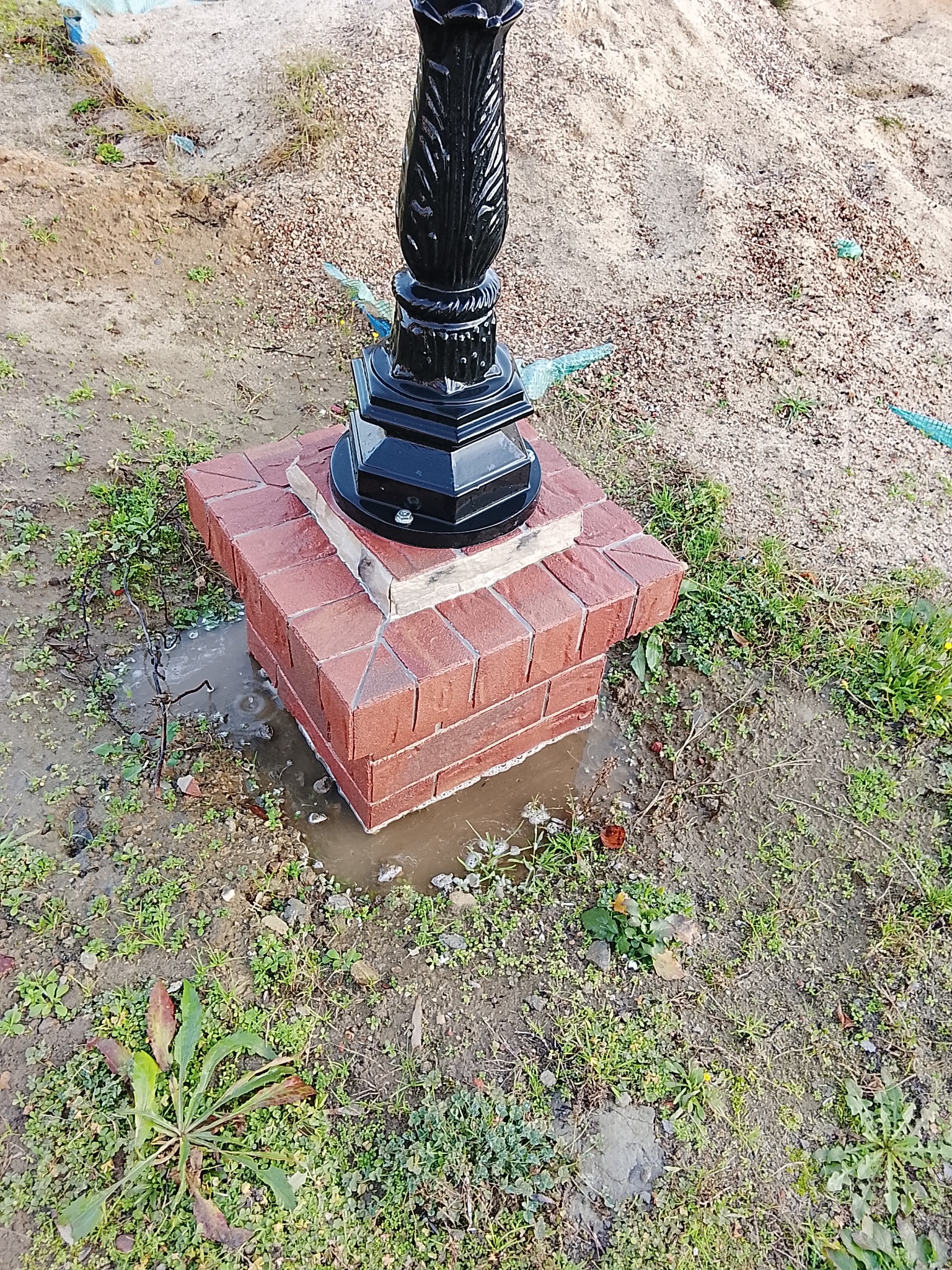 Czarna, ozdobna lampa ogrodowa na cokole z czerwonej cegły, otoczona ziemią i zielenią. Widoczne ślady wilgoci wokół podstawy. Ujęcie z bliska, podkreślające detale.