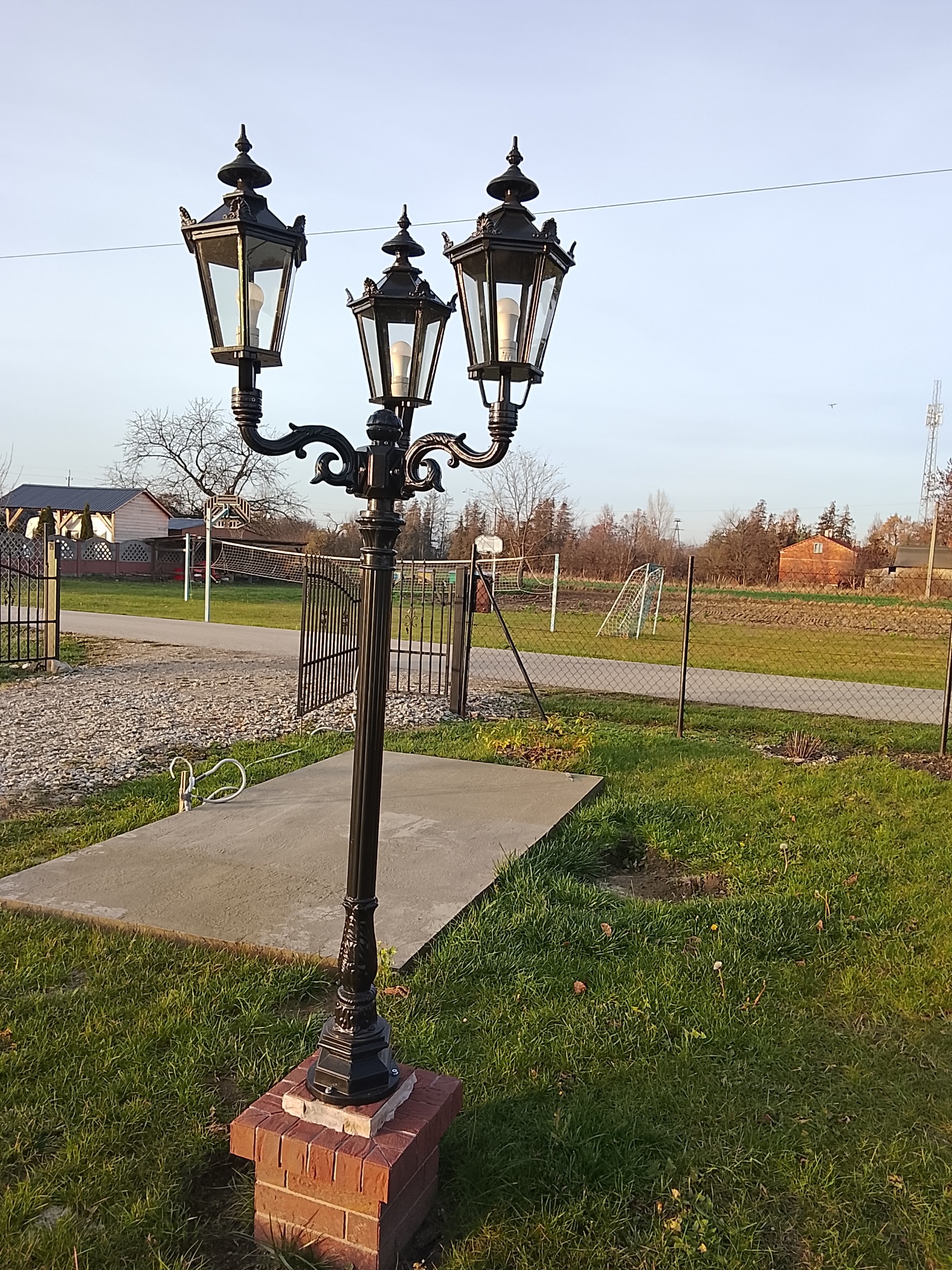Ozdobna, czarna, trójramienna latarnia parkowa na ceglanym cokole, oświetlająca teren zielony z widocznymi elementami infrastruktury sportowej w tle.