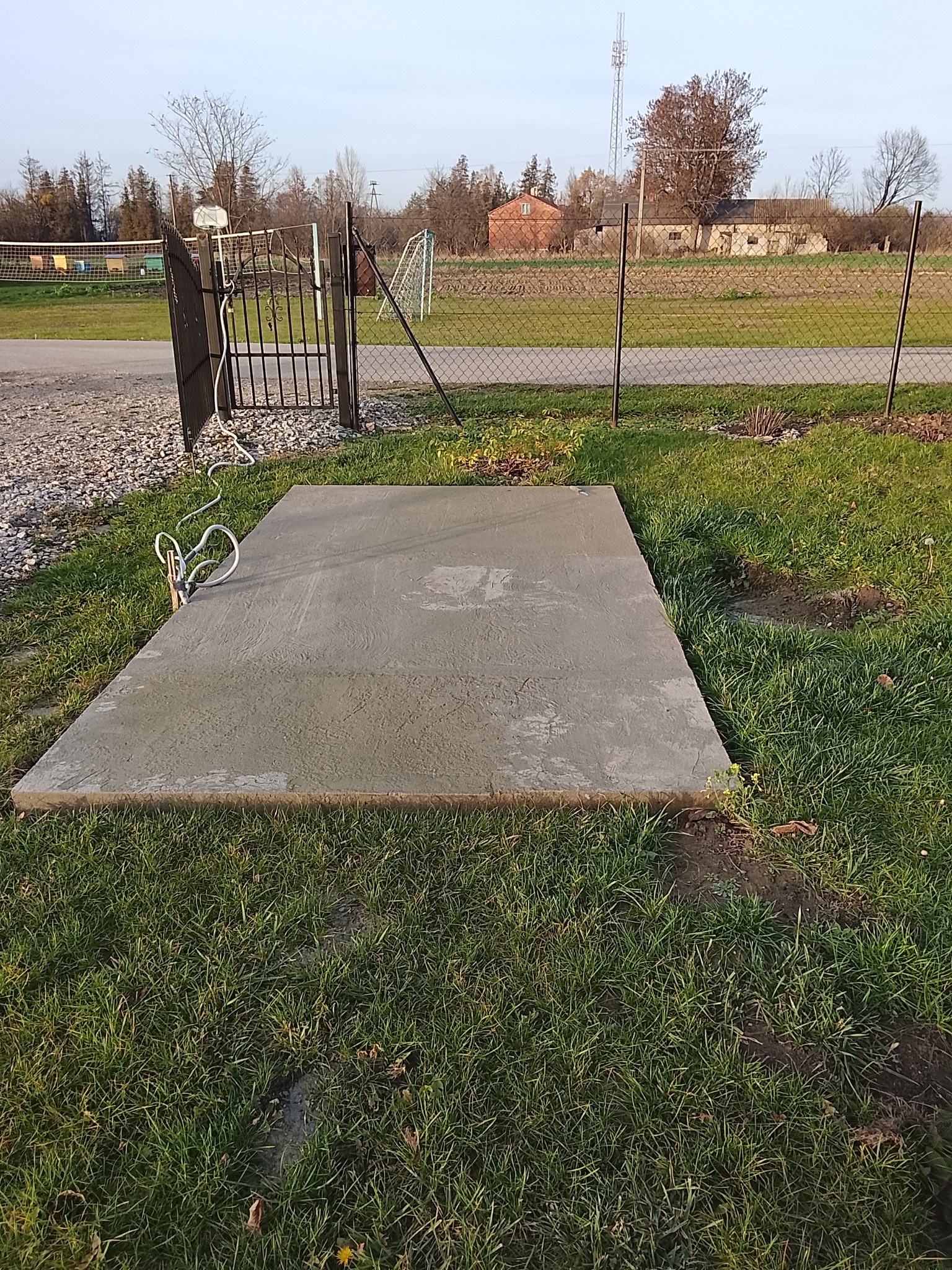 Świeżo wylana, prostokątna płyta betonowa na trawie, z widocznymi śladami po narzędziach, obok furtki z metalową siatką i widokiem na wiejskie zabudowania w tle.