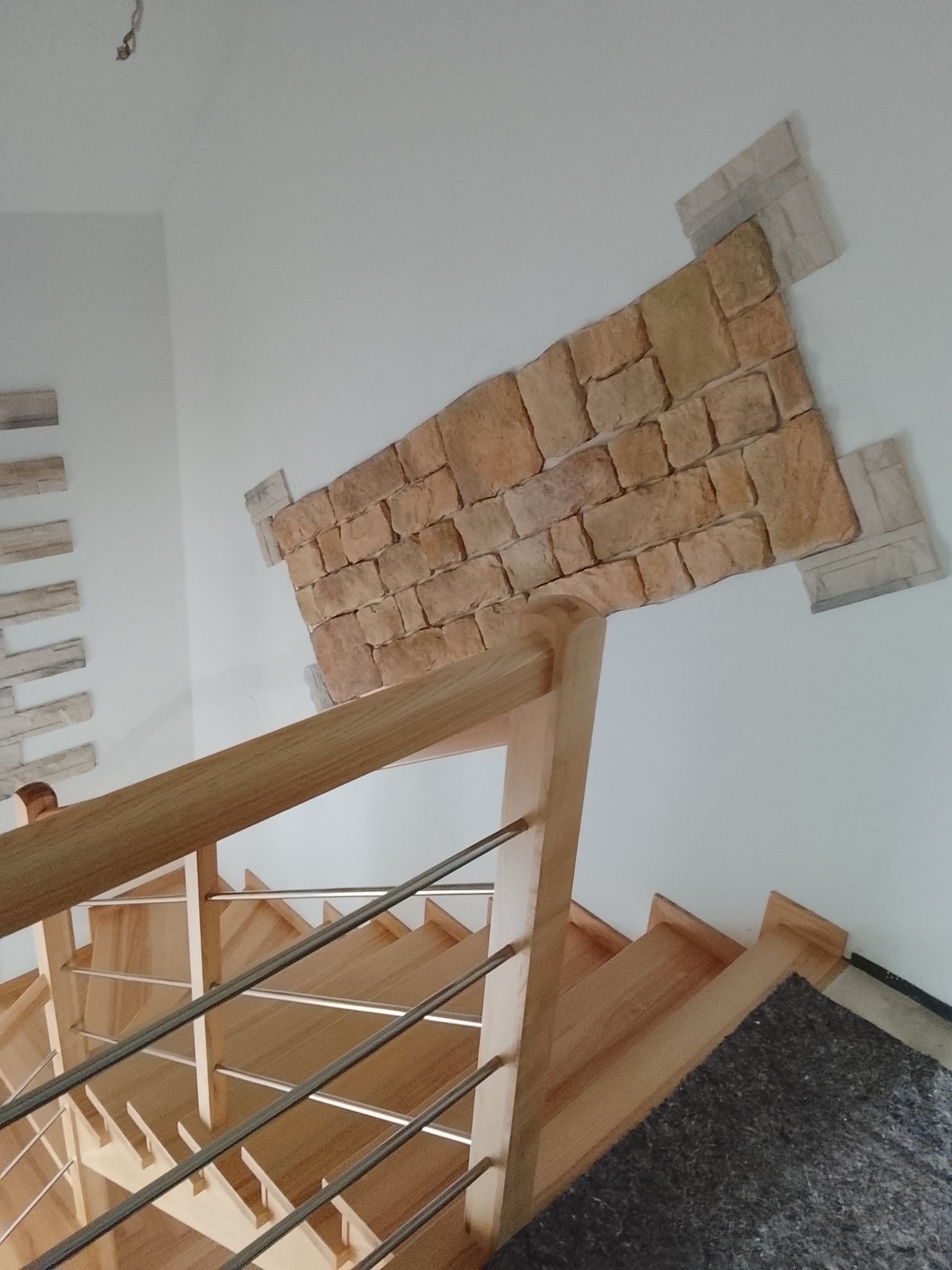 Wykończenie ściany przy schodach imitacją kamienia w ciepłych odcieniach, z drewnianą balustradą i metalowymi elementami. Ujęcie z góry, widoczne schody.