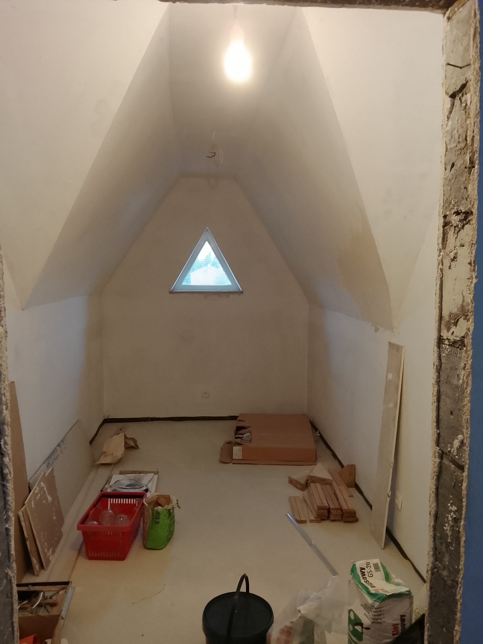 Poddasze w trakcie remontu: surowe ściany, trójkątne okno, materiały budowlane i narzędzia. Widok na wnętrze przez surową futrynę.