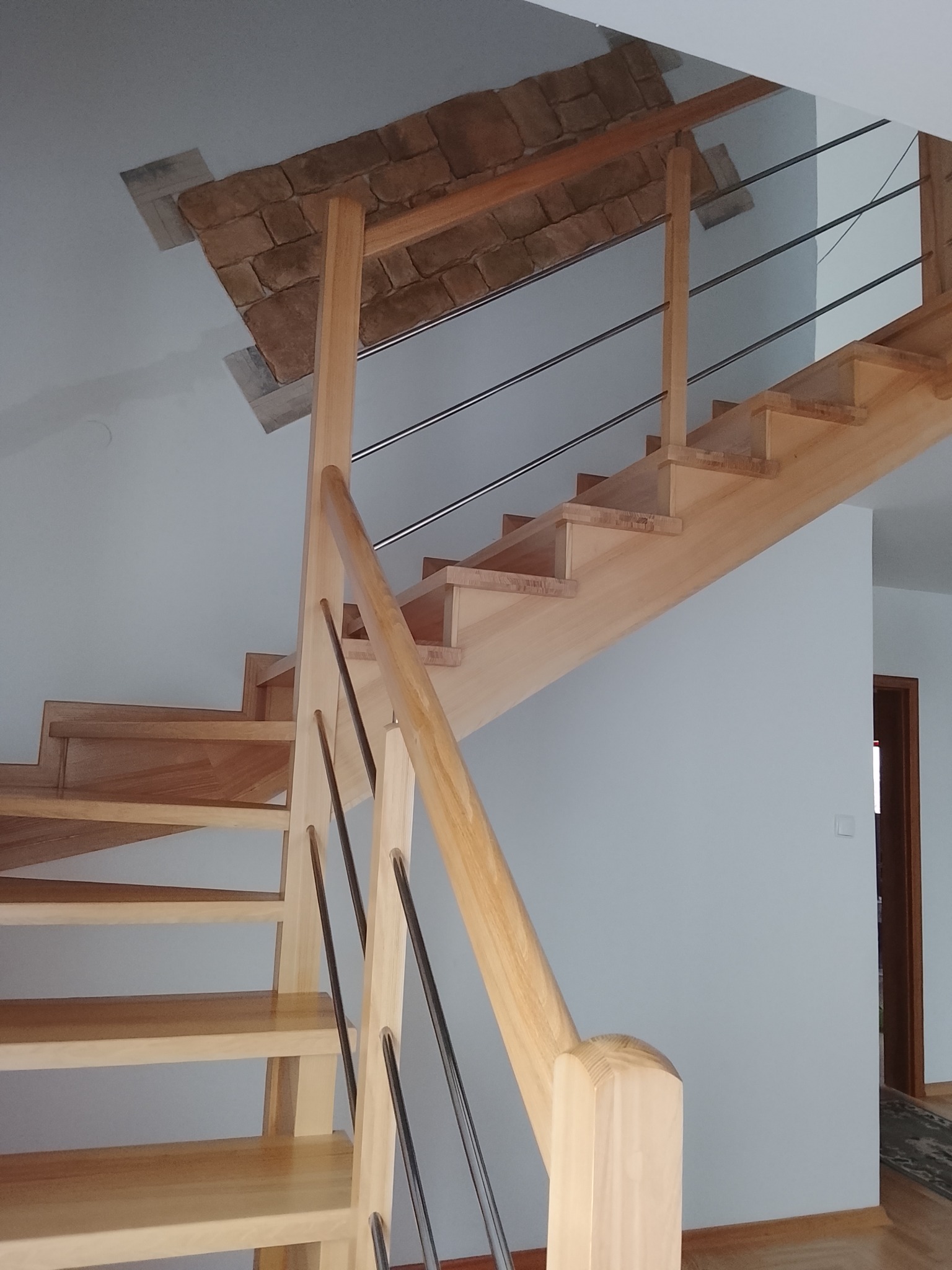 Jasne, drewniane schody z metalowymi barierkami w nowoczesnym wnętrzu. Fragment dekoracyjnej, kamiennej obudowy sufitu nad schodami. Widok z dołu, perspektywa.