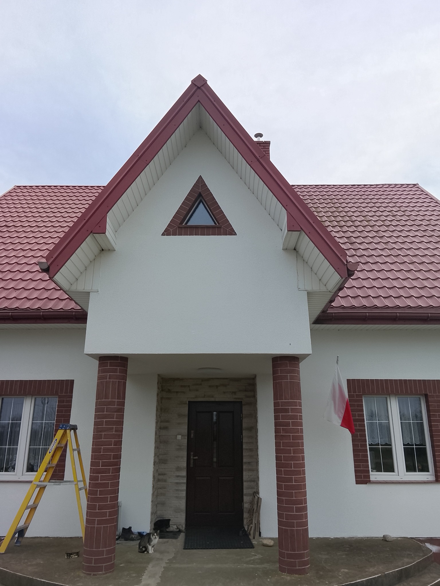 Biały dom z czerwoną dachówką i ceglanymi detalami, flaga Polski, drabina oparta o ścianę, kot siedzący przed wejściem. Widok z dołu, pochmurne niebo.
