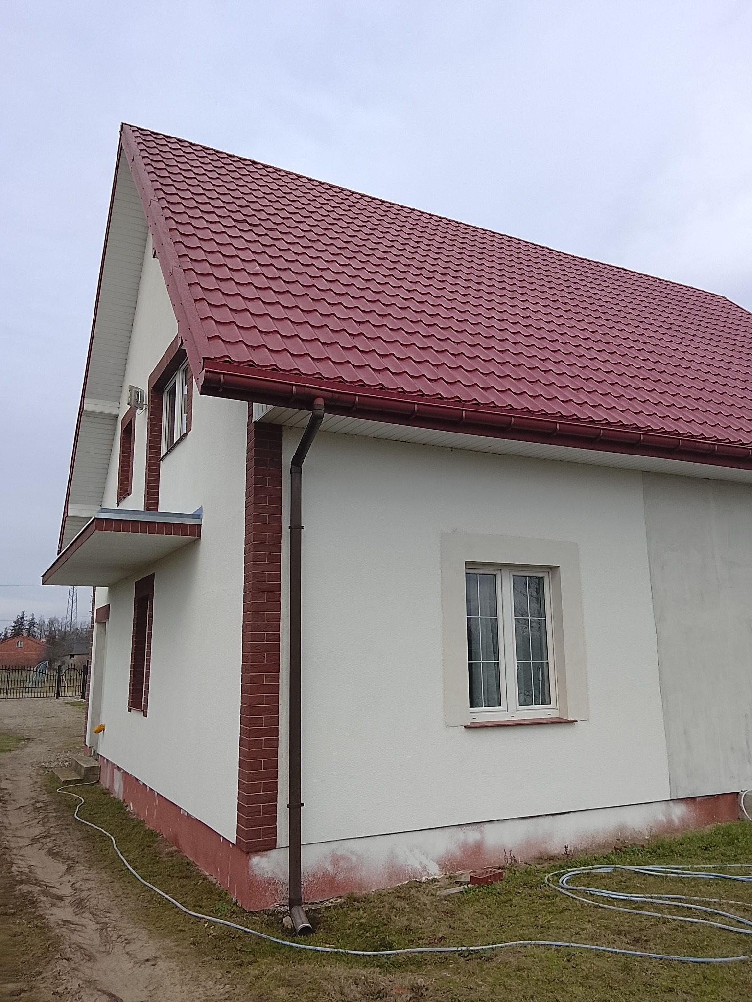 Boczny widok domu z czerwoną dachówką i białymi ścianami, obramowanymi cegłą. Widoczne okna i rynna. Tło stanowi pochmurne niebo i fragment otoczenia.