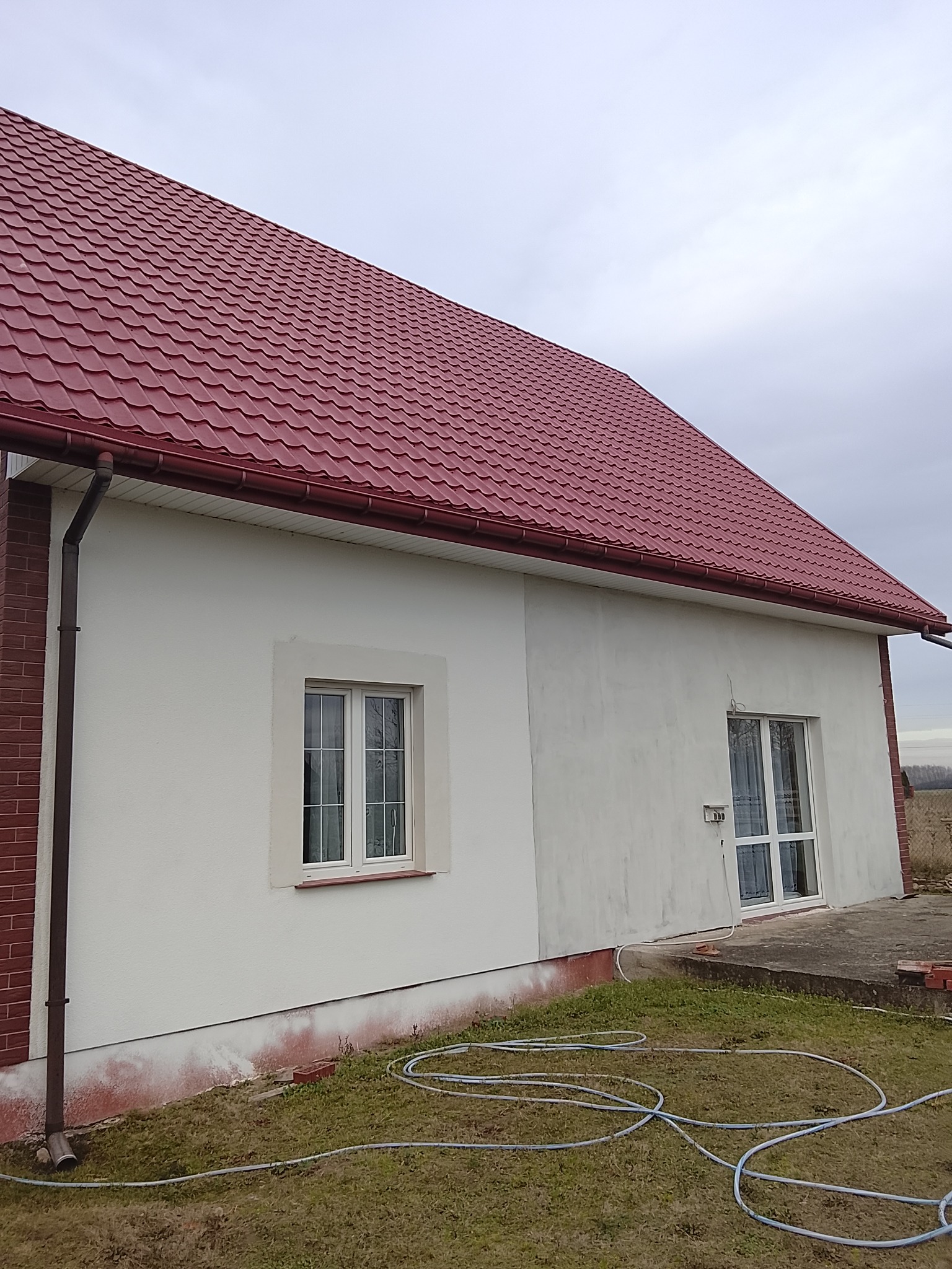 Wykończony dom z czerwoną dachówką i białą elewacją. Widoczne okna z białymi ramami, rynny i fragment muru z cegły. Na trawie leży zwinięty wąż ogrodowy.