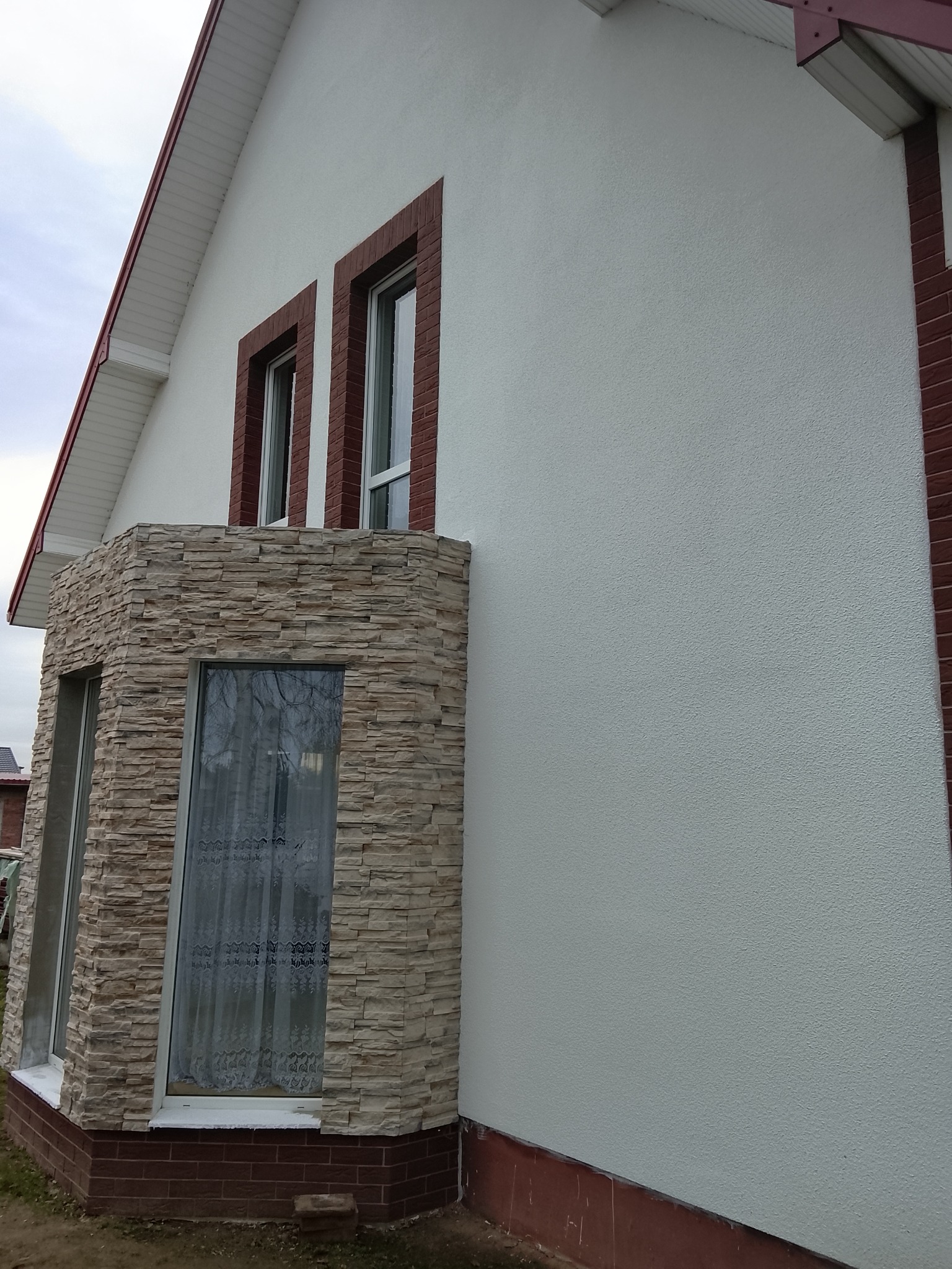 Elewacja domu z oknami obramowanymi czerwoną cegłą i wykuszem wykończonym kamieniem dekoracyjnym. Biały tynk strukturalny na ścianie. Ujęcie z dołu, lekki kąt.