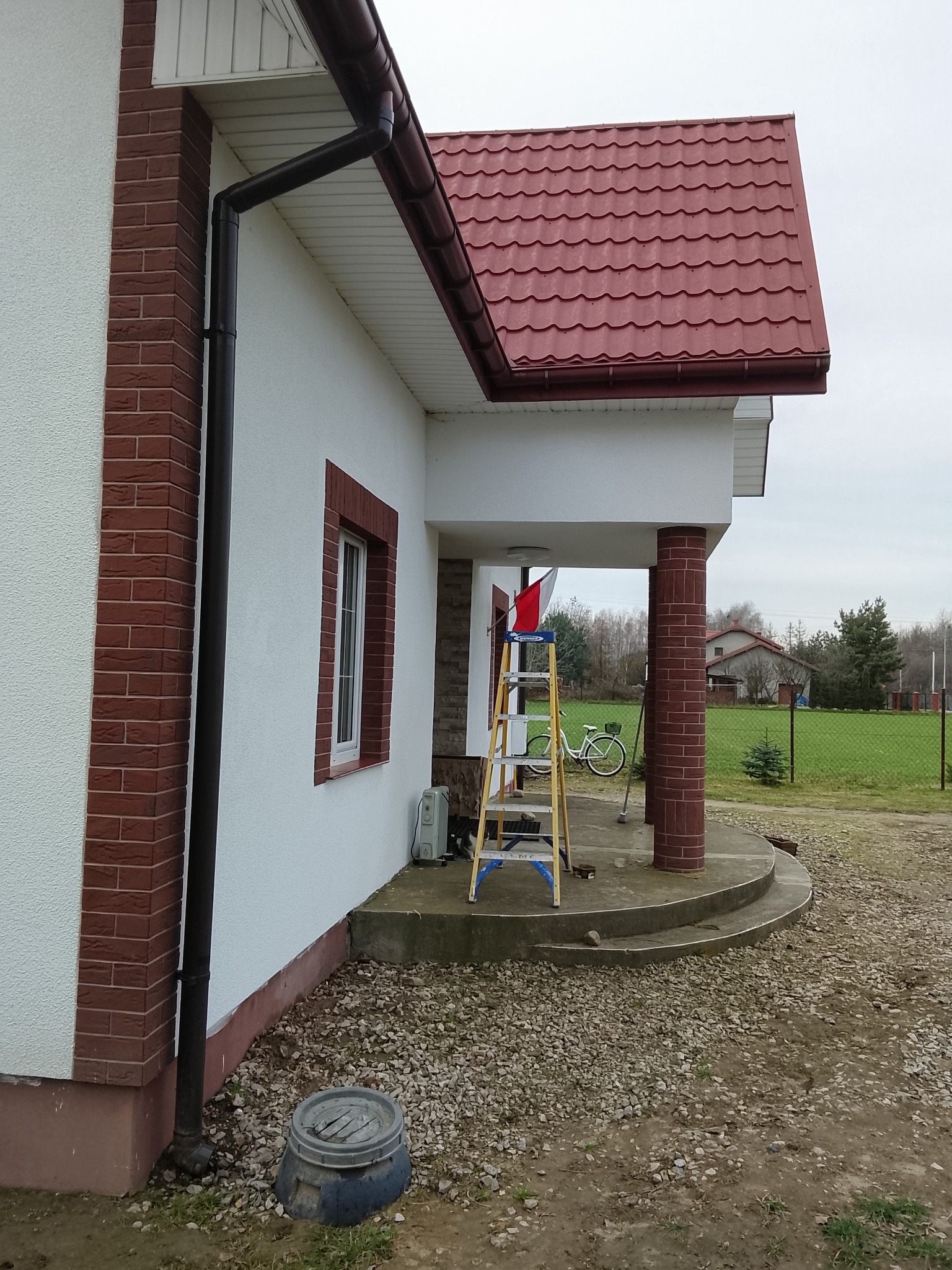 Dom jednorodzinny z elewacją w trakcie prac wykończeniowych, widoczna drabina i fragment elewacji z cegły klinkierowej. Dach pokryty czerwoną blachodachówką.