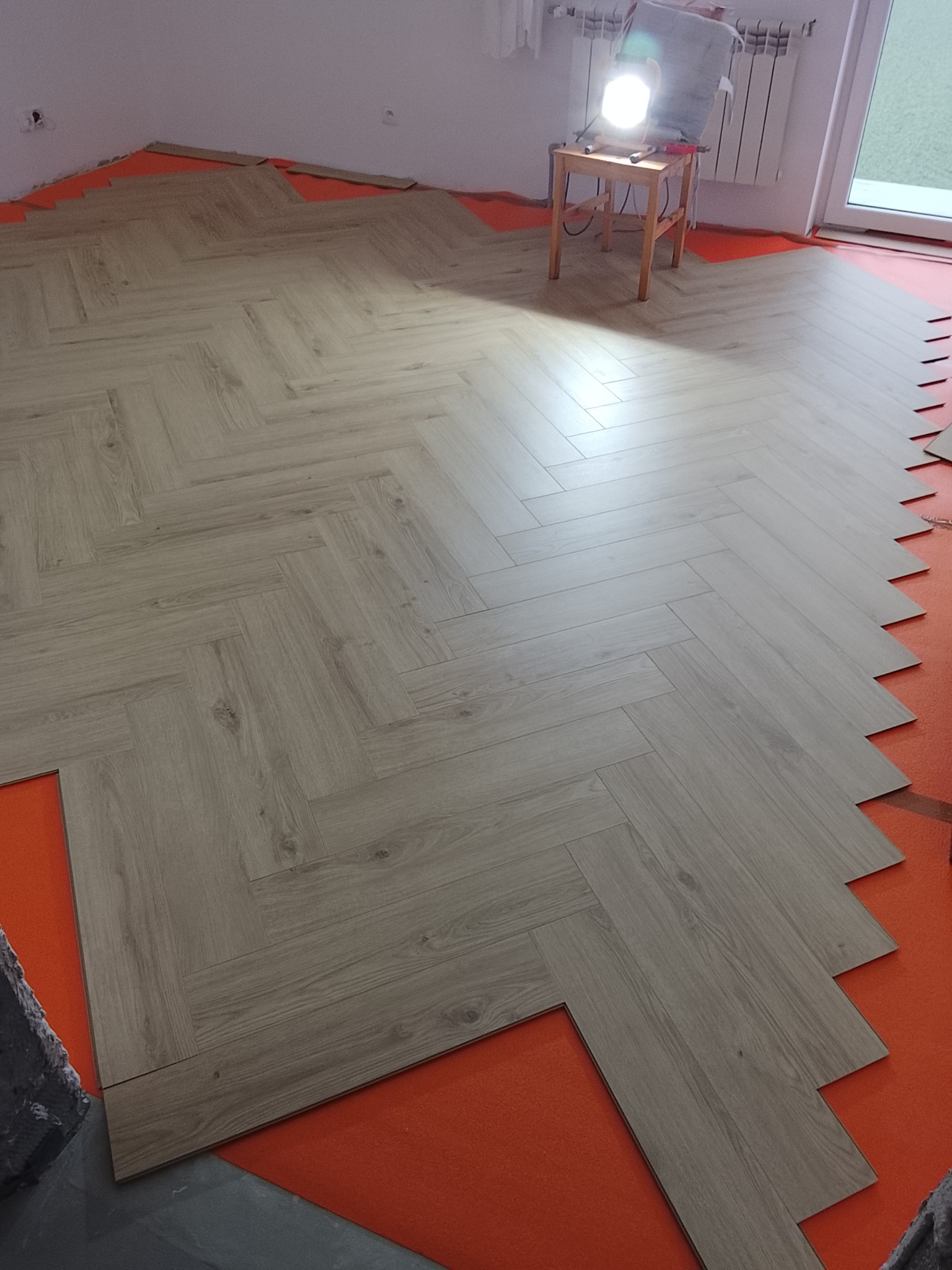 Układanie paneli podłogowych w jodełkę na pomarańczowym podkładzie. Widoczna lampa warsztatowa i grzejnik. Częściowo ułożona podłoga w jasnym odcieniu drewna.