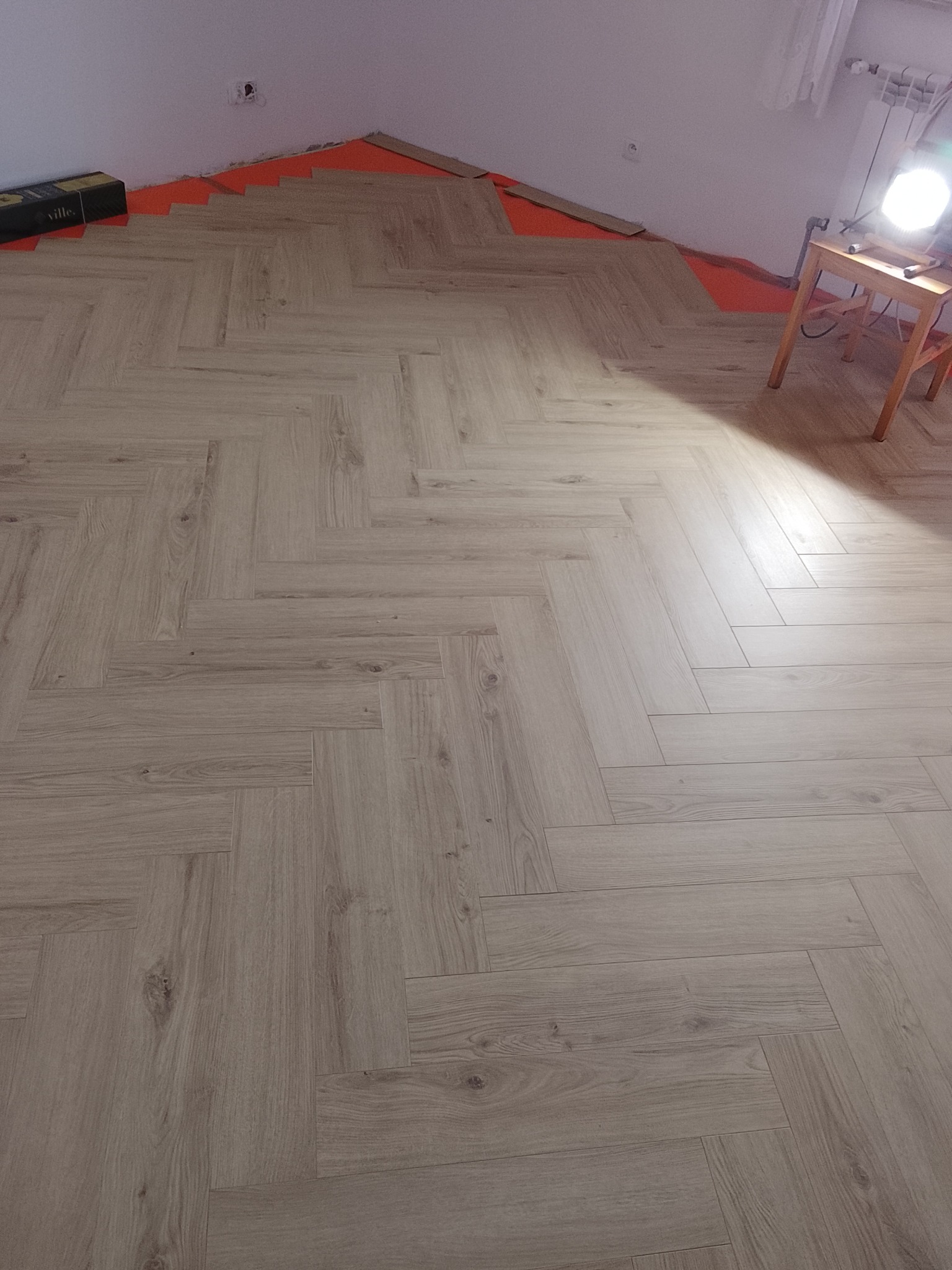 Układanie parkietu w jodełkę: widok z góry na jasne panele podłogowe, częściowo ułożone na pomarańczowej macie izolacyjnej, z lampą warsztatową w rogu.