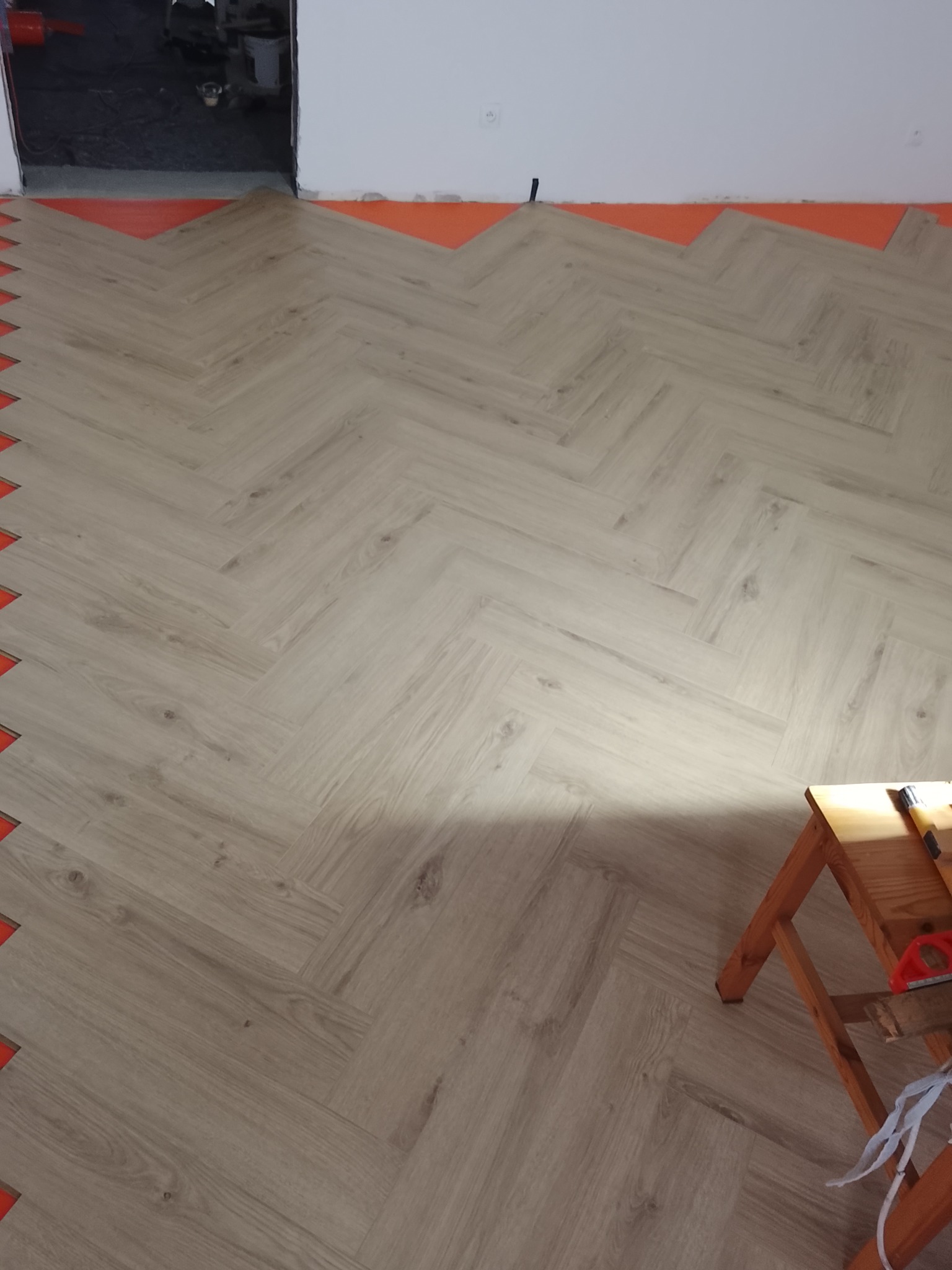 Układanie paneli podłogowych w jodełkę, widoczne pomarańczowe podłoże i drewniany taboret z narzędziami w rogu. Częściowo wykończona podłoga.